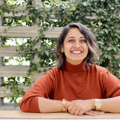 Dr Jaina Chauhan smiles at the camera in front of background of greenery. She has medium length dark hair and is wearing a burnt orange long sleeve top.