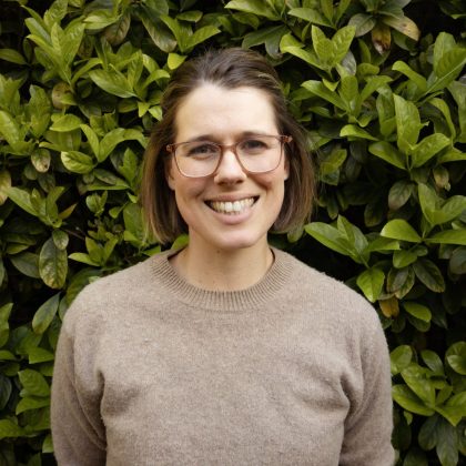 Dr Alicia Kaczmark smiles at the camera in front of a background of green bushes. She has short dark hair and is wearing glasses.