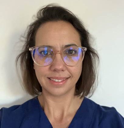 Dr Liesl Larsen smiles at the camera in front of a white wall. She has short dark hair and is wearing glasses and a navy scrub top