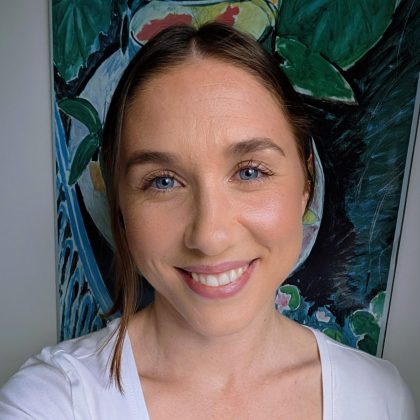 Dr Ellen Robinson smiles at the camera. She is wearing an all white outfit and standing in front of a colourful painting